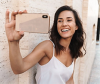 A smiling woman with dark brown tousled hair leaning against a wall and taking a selfie 