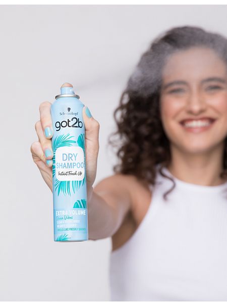 Dark-haired woman with curly hair spraying Got2b Dry Shampoo Ocean Vibes at the camera.