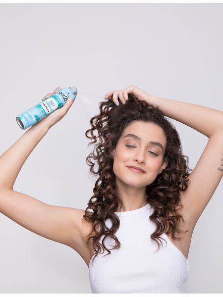 Dark-haired woman with curly hair spraying Got2b Dry Shampoo Ocean Vibes onto her locks.