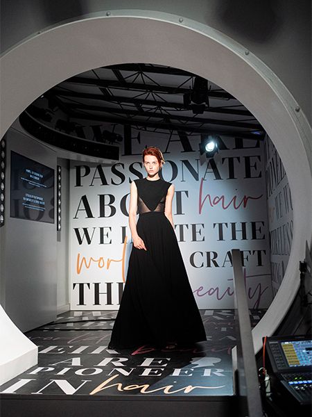 Red haired model on the runway wearing a black dress, behind her a white wall with black writing. The floor is black with white writing. She is walking through a white round arch.