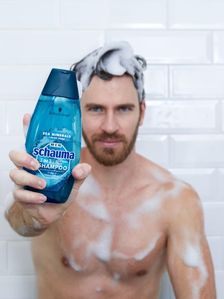 Man soaped up in the shower, holding a bottle of Schauma 3 in 1 Shampoo For Men