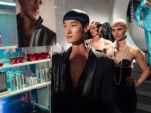 Three models photographed from the side, looking straight-ahead, each with a modern hairstyle and wearing black clothing, next to them a shelf full of hair products.