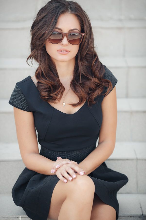 Femme brune dans une robe noire, assise sur des marches et portant des lunettes de soleil.