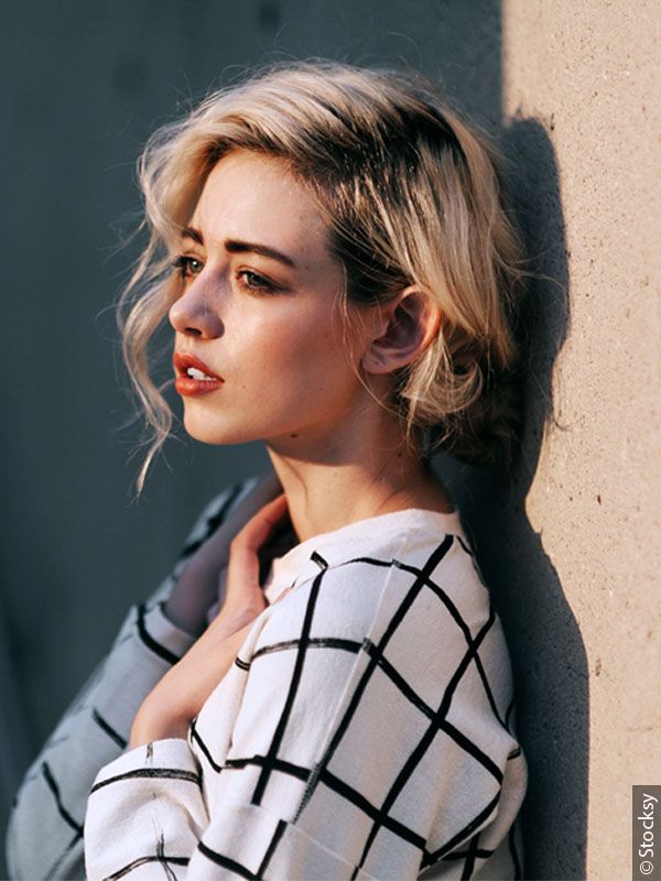 Woman with blond faux bob wearing a black and white top and leaning against a wall  
