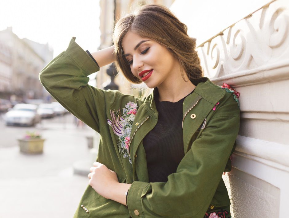 Femme brune avec un balayage blond et une parka kaki se tenant contre un mur