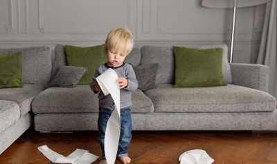 little boy standing in a living room in front of a sofa looking at a roll of toilet paper in his hands