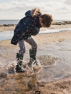Jacke waschen, Junge mit  dunkler warmer jacke und  gummistiefeln springt in pfütze am  meer