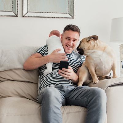 Mężczyzna siedzi na kanapie, trzyma w rękach niemowlaka, obok siedzi pies.