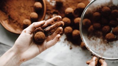 Kakao ile kirlenmiş bir el taze hazırlanmış bir çikolatalı pralin tutuyor, arka plandaki kasede ve masada ise bulanık pralinler duruyor.