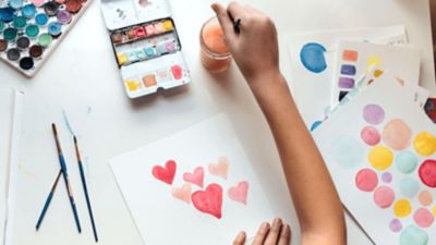 Χέρια παιδιών που χρησιμοποιούν ακουαρέλες για να ζωγραφίσουν κόκκινες καρδιές σε ένα λευκό φύλλο χαρτιού.