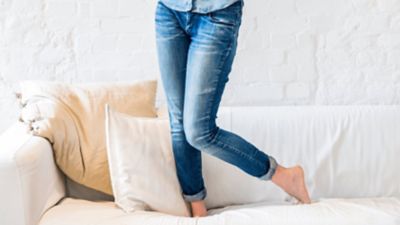 Μια γυναίκα που φορά τζιν στέκεται σε έναν μαλακό καναπέ, μόνο τα πόδια της είναι ορατά.