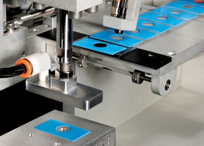 Close up photo of blue rectangular sil pads with circular hole in center being manufactured and placed on tape by automation equipment