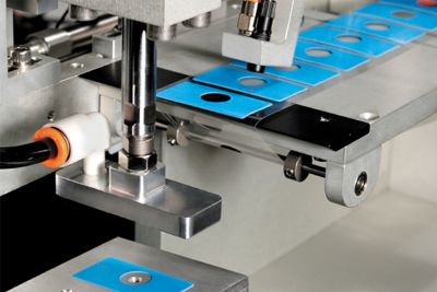 Close up photo of blue rectangular sil pads with circular hole in center being manufactured and placed on tape by automation equipment