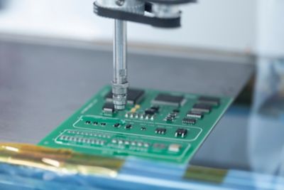 Photo of nozzle dispensing conformal coating onto a blue green printed circuit  for protection, waterproofing and improved reliability