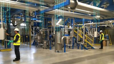 Photo of two employees working with digital interface near stairs tanks and pipes inside the montornes del valles spain smart adhesives manufacturing facility embellished with binary code streaming from above to represent digitalization of the factory