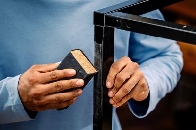 A hand works on a metal frame with sandpaper, while the other hand holds the frame in place.