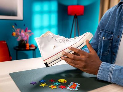 Two hands holding a sneaker with a tear. Beneath are colourful patches on a table.