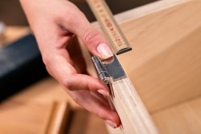 Uma mão medindo e posicionando uma dobradiça de metal sobre uma peça de madeira.