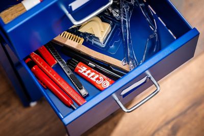 Open drawer with safety goggles, a folding rule, pencils, a cutter knife, a wire brush, and glue.