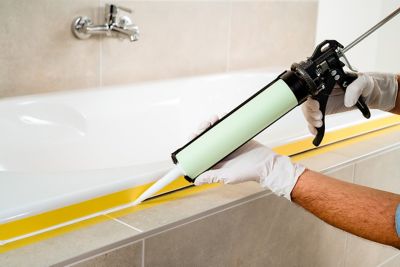 Hands seal the edge of a bathtub with a caulking gun, between two strips of masking tape.