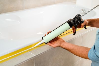 A man with a caulking gun applies some product to seal a bathtub. The edges are taped.