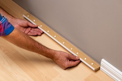 One hand holds a baseboard horizontally to the wall, and on the backside is a line with glue dots.