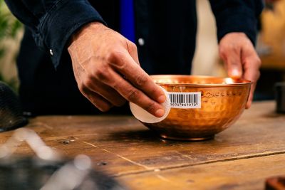 A hand presses a cotton pad onto the stick-on barcode label of a metal bowl.