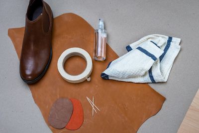 A leather shoe, three toothpicks, two patches, a bottle with transparent liquid, a rag and tape on a leather surface.