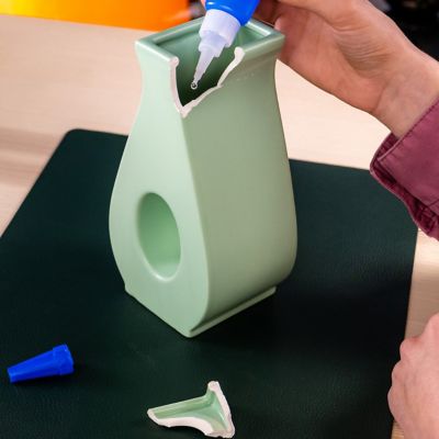 A person squeezing a drop of liquid super glue onto a broken vase, the broken piece lays next to it.