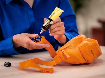 Eine Hand trägt Klebstoff auf den gebrochenen Plastikschnappverschluss einer orangefarbenen Tasche auf.