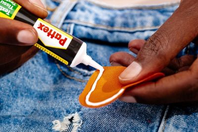 Hand trägt Kleber auf orangenem Flicken über einer Jeans mit Loch auf. 