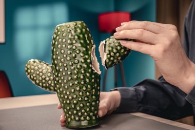 Alt text: Grøn vase i form af en kaktus. Den ene arm er knækket af og holdes op mod vasen.