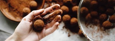 Kakao ile kirlenmiş bir el taze hazırlanmış bir çikolatalı pralin tutuyor, arka plandaki kasede ve masada ise bulanık pralinler duruyor.