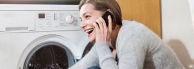 Besijuokianti moteris tupi priešais skalbyklę ir kalba telefonu.