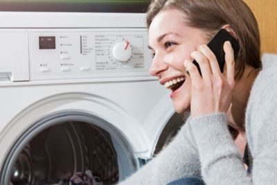 Une femme souriant au téléphone assise devant sa machine à lavée 