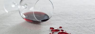 Prevrnjen kozarec z rdečim vinom, na beli preprogi kapljice vina.