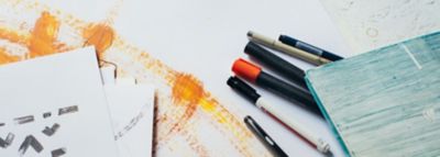 Baltame fone - įvairūs pieštukai; per vidurį nubrėžtos geltonos, oranžinės ir rudos spalvos linijos. 