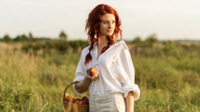 Junge rothaarige Frau mit weißem Leinenhemd und beigefarbener Hose steht in hohem Gras mit einem Korb voller Pfirsiche und einem Pfirsich in der Hand