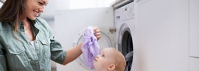 Mati smeje gleda svojega dojenčka medtem ko jemlje perilo iz pomivalnega stroja.