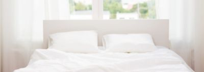 Un pat cu cearceafuri albe de in, poziționat în fața unui geam. 