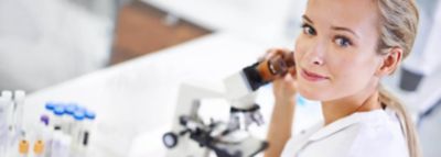 Eine Frau in weißer Laborkleidung schaut vom Mikroskop auf und blickt über ihre linke Schulter in die Kamera.