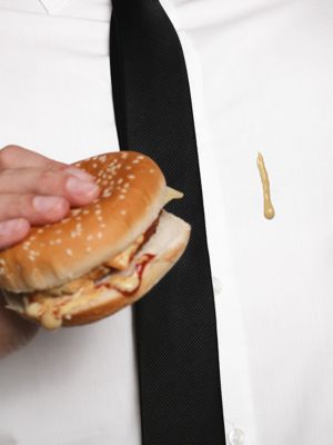 Mann Hemd mit Senffleck und schwarzer Krawatte hält Burger in der Hand 
