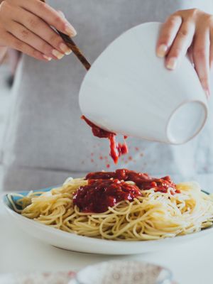 Frau schüttet rote Sauce aus weißer Schüssel auf Teller mit Spaghetti 