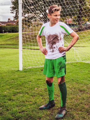 Junge in weiß-grünem Fußball Trikot voller Erdflecken steht vor einem Fußballtor auf dem Rasen