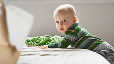 Blondes Baby liegt in einem grün-gestreiften Longsleeve und einer grauen Hose auf einem Bett.