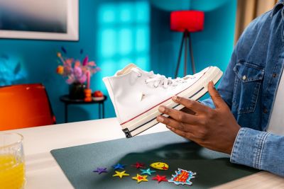 Two hands holding a sneaker with a tear. Beneath are colourful patches on a table.