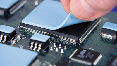 Photo of blue  thermal gap pad being placed on top of an electronic component on a pcb with other blue thermal gap pads on other components elsewhere on the pcb assembly